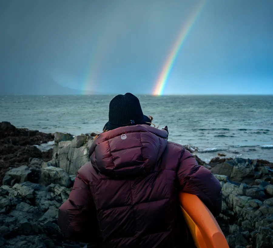 Sailor in a down parka on a rocky shore, rainbow over the sea